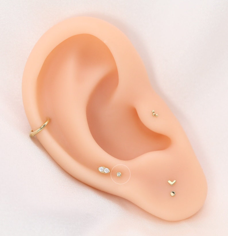 May include: A display of various gold earrings on a mannequin ear. The earrings include a small hoop, a trio of small balls, a small stud, a double-stone oval, a small round stud, and a chevron-shaped earring.