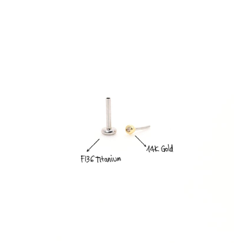 May include: Two pieces of body jewelry. One is a silver-colored F136 titanium labret stud. The other is a 14K gold labret stud with a small, clear gem. Both are on a white background.
