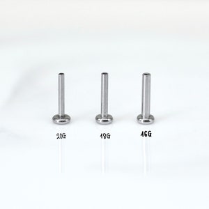 May include: Three silver-colored body jewelry studs with flat, round bases and cylindrical posts. The studs are labeled with sizes: 20G, 18G, and 16G. The background is plain white.