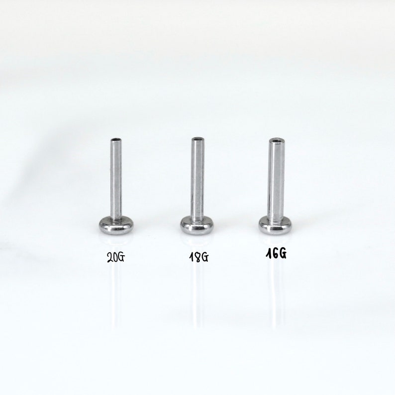 May include: Three silver-colored body jewelry studs with flat, round bases and cylindrical posts. The studs are labeled with sizes: 20G, 18G, and 16G. The background is plain white.