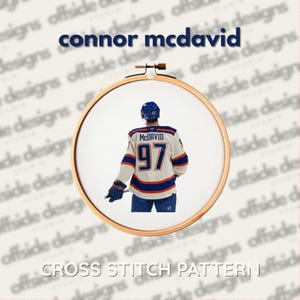 Op de afbeelding: Een kruissteekpatroon met een hockeyspeler in een trui met de naam "McDavid" en het nummer "97". Het ontwerp is omlijst in een houten borduurring, met de tekst "Connor McDavid" en "Cross Stitch Pattern".