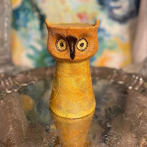 May include: A ceramic owl-shaped vase with a textured, yellow and orange finish. The owl has large, round eyes and a dark brown beak. The vase is on a silver tray, and the background is a colorful, blurred pattern.