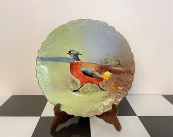 May include: A decorative plate featuring a pheasant illustration. The plate has a scalloped edge with gold trim. The pheasant is depicted with red, brown, and blue plumage against a green and blue background. It is displayed on a wooden stand.