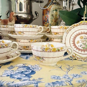 Könnte beinhalten: Ein Set vintage Teetassen und Untertassen mit einem floralen Muster in Braun-, Gelb- und Weißtönen. Die Tassen und Untertassen haben einen gewellten Rand und sind auf einer gelben Tischdecke mit einem blauen Blumenmuster angeordnet.