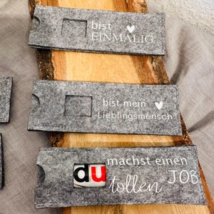 Puede incluir: Tres porta tarjetas de regalo de fieltro gris con texto blanco. El primer porta tarjetas dice "bist einmalig" con un coraz&oacute;n. El segundo porta tarjetas dice "bist mein Lieblingsmensch" con un coraz&oacute;n. El tercer porta tarjetas dice "du machst einen tollen JOB".