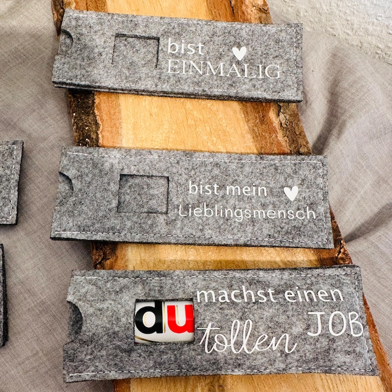 Puede incluir: Tres porta tarjetas de regalo de fieltro gris con texto blanco. El primer porta tarjetas dice "bist einmalig" con un coraz&oacute;n. El segundo porta tarjetas dice "bist mein Lieblingsmensch" con un coraz&oacute;n. El tercer porta tarjetas dice "du machst einen tollen JOB".