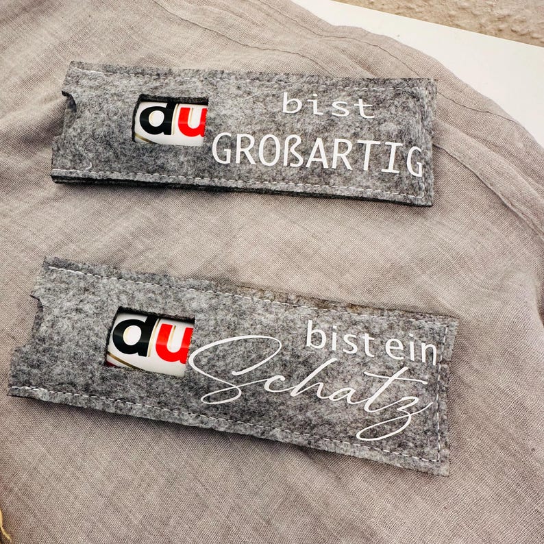 Puede incluir: Dos bolsas de regalo de fieltro gris con letras blancas. La bolsa superior dice "Du bist gro&szlig;artig" y la bolsa inferior dice "Du bist ein Schatz".