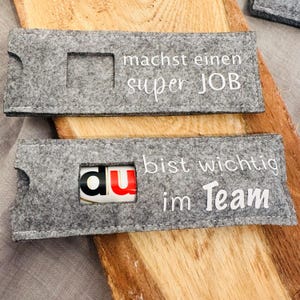 Puede incluir: Dos bolsas de regalo de fieltro gris con texto blanco. La bolsa de arriba dice "machst einen super JOB". La bolsa de abajo dice "bist wichtig im Team" y tiene una peque&ntilde;a barra de chocolate dentro.
