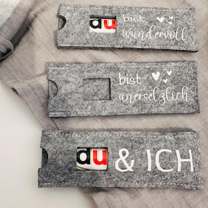 Puede incluir: Tres bolsas de regalo de fieltro gris con texto blanco y corazones. Las bolsas tienen una peque&ntilde;a ventana para insertar un peque&ntilde;o regalo. El texto en las bolsas dice "Du bist wundervoll", "Du bist unersetzlich" y "Du & ICH".