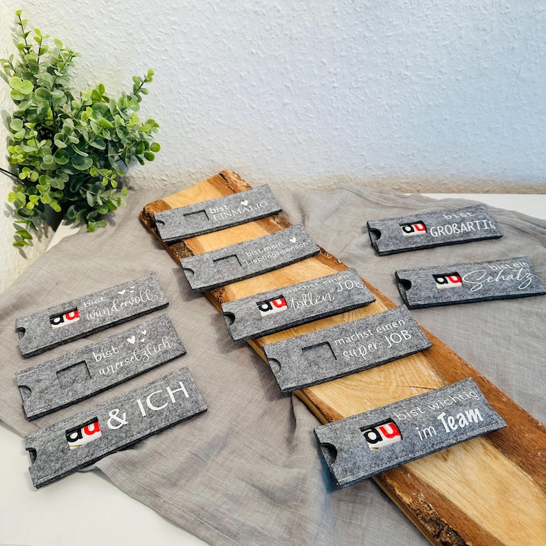 Puede incluir: Un conjunto de diez cajas de regalo de fieltro gris con texto blanco y un logotipo rojo. El texto en las cajas incluye "Eres incre&iacute;ble", "Eres maravilloso", "Eres una gran persona", "Haces un gran trabajo", "Haces un gran trabajo", "Haces un gran trabajo", "Haces un gran trabajo", "Haces un gran trabajo", "Haces un gran trabajo", "Haces un gran trabajo".