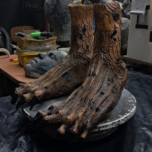 Puede incluir: Un par de pies detallados, marrones, con forma de árbol, con piel texturizada similar a la corteza y largas garras oscuras. Los pies están colocados sobre una superficie redonda y gris, lo que sugiere un tema teatral o de diseño de vestuario.