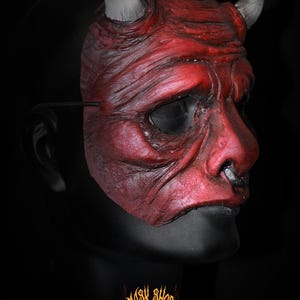 Puede incluir: Una m&aacute;scara de diablo roja con cuernos blancos y un aro en la nariz negro. La m&aacute;scara tiene una superficie texturizada y est&aacute; sobre un fondo negro. El logo de Mask Shop es visible en la parte inferior.