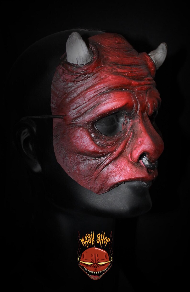 Puede incluir: Una m&aacute;scara de diablo roja con cuernos blancos y un aro en la nariz negro. La m&aacute;scara tiene una superficie texturizada y est&aacute; sobre un fondo negro. El logo de Mask Shop es visible en la parte inferior.