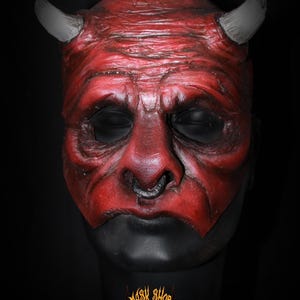 Puede incluir: Una m&aacute;scara de diablo roja con cuernos grises y una base negra. La m&aacute;scara tiene una apariencia envejecida detallada con arrugas y un piercing. El logotipo de Mask Shop es visible en la base.