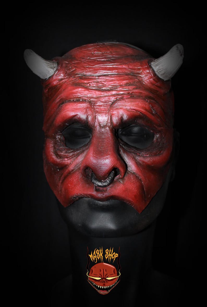 Puede incluir: Una m&aacute;scara de diablo roja con cuernos grises y una base negra. La m&aacute;scara tiene una apariencia envejecida detallada con arrugas y un piercing. El logotipo de Mask Shop es visible en la base.