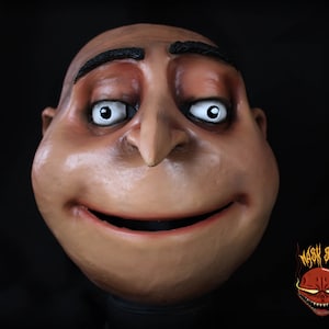 Puede incluir: Un primer plano de una máscara realista de Gru de las películas de Gru, mi villano favorito. La máscara está hecha de látex y presenta una escultura detallada de la cara de Gru, incluyendo su cabeza calva característica, cejas y bigote. La máscara tiene una expresión neutral con una leve sonrisa.