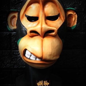 May include: A cartoon monkey mask in orange and black, with green detailing. The mask features a wide grin and closed eyes. The mask is displayed on a black stand, with the text "MASK SHOP" and a skull logo. A striking design.