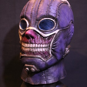 Pu&ograve; includere: Una maschera viola e argento con una texture cucita e grandi occhiali. La maschera ha un ampio sorriso pieno di denti e un naso rosa.