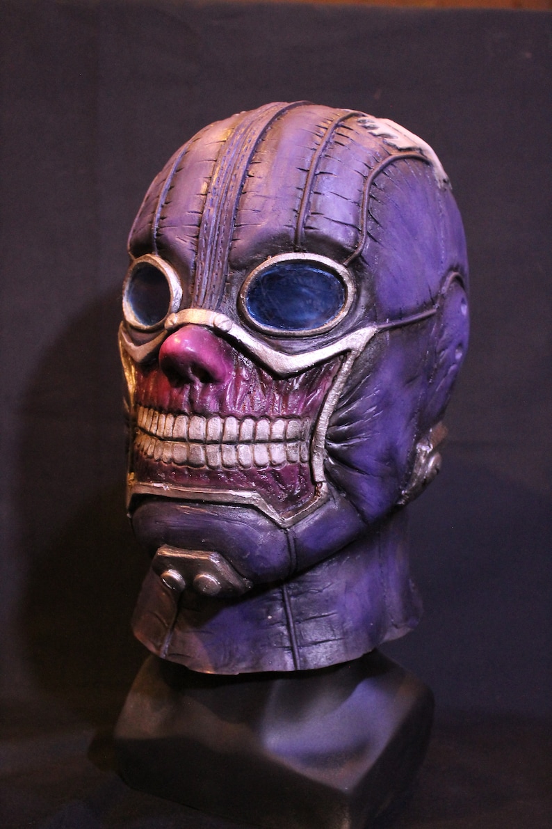 Pu&ograve; includere: Una maschera viola e argento con una texture cucita e grandi occhiali. La maschera ha un ampio sorriso pieno di denti e un naso rosa.
