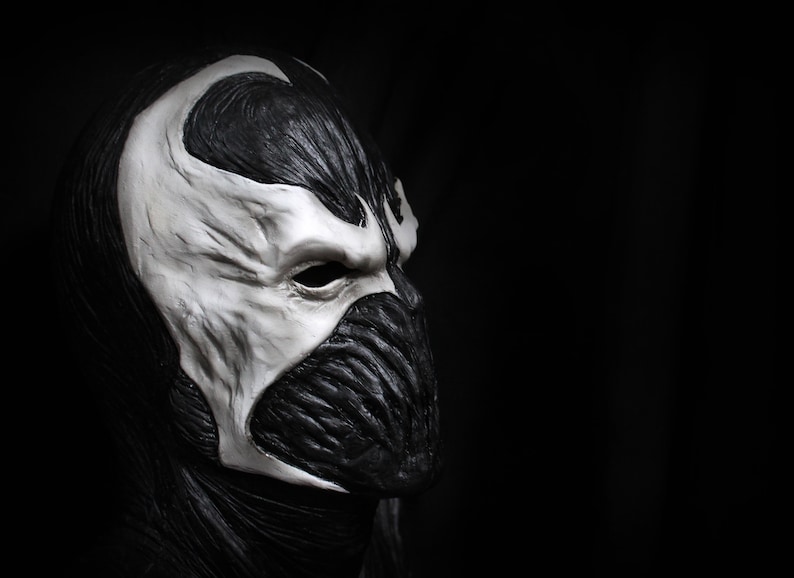 May include: A black and white mask with a detailed design. The mask has a sharp, angular shape and a single eye hole. The mask is made of a smooth, textured material.