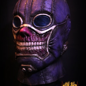 Pu&ograve; includere: Una maschera di teschio viola e argento con un design cucito e occhiali. La maschera ha un sorriso dettagliato e dentato e un naso rosa.