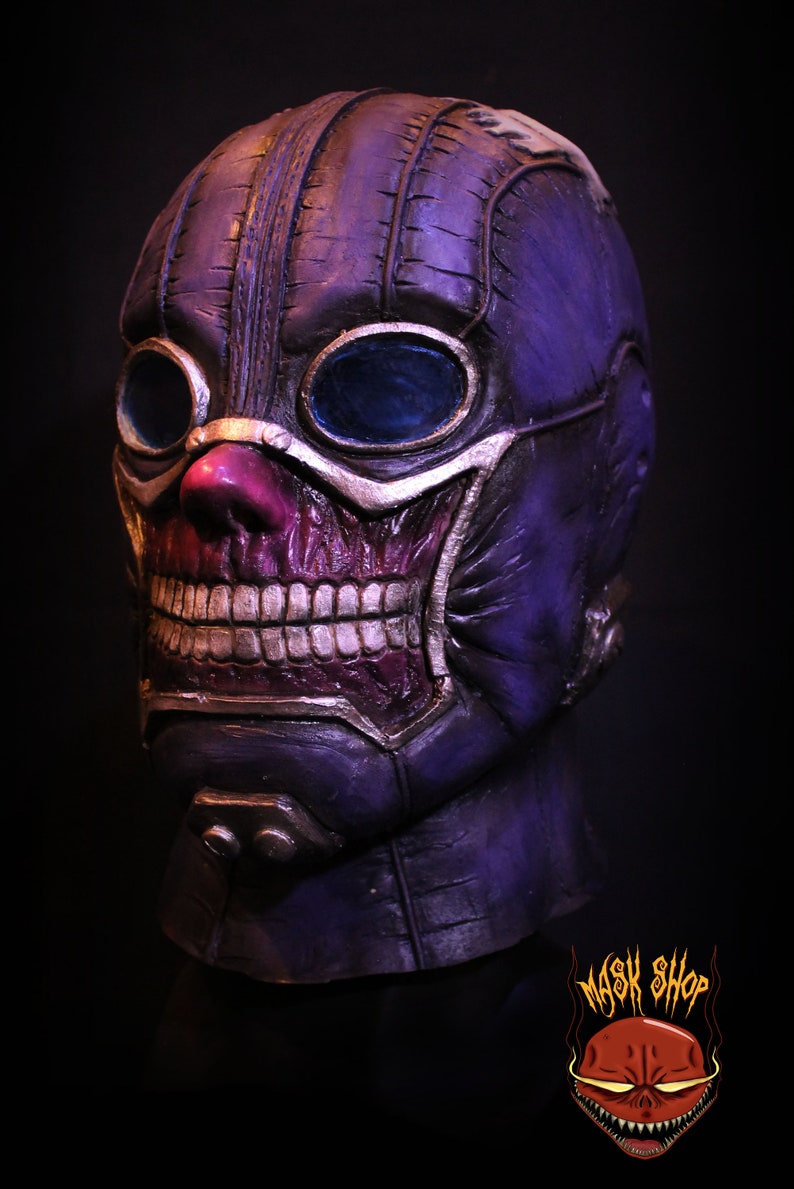 Pu&ograve; includere: Una maschera di teschio viola e argento con un design cucito e occhiali. La maschera ha un sorriso dettagliato e dentato e un naso rosa.