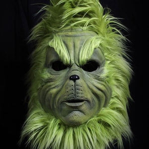 Puede incluir: Una máscara verde con una expresión gruñona, con una cara texturizada y una melena verde y peluda. La máscara tiene cuencas oscuras y una pequeña nariz negra. La parte inferior de la máscara tiene el texto "MASK SHOP".