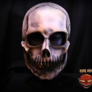Puede incluir: Una máscara de calavera detallada con un acabado blanquecino desgastado y sombreado oscuro. La máscara presenta un diseño esquelético realista con dientes y cuencas de los ojos visibles. La máscara tiene un logotipo en la esquina inferior derecha que dice "MASK SHOP".