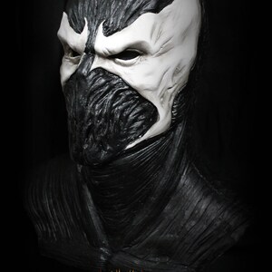 May include: A black and white detailed mask of a character with a large, open mouth and sharp teeth. The mask is made of a textured material and has a realistic appearance. The mask is on a black background.