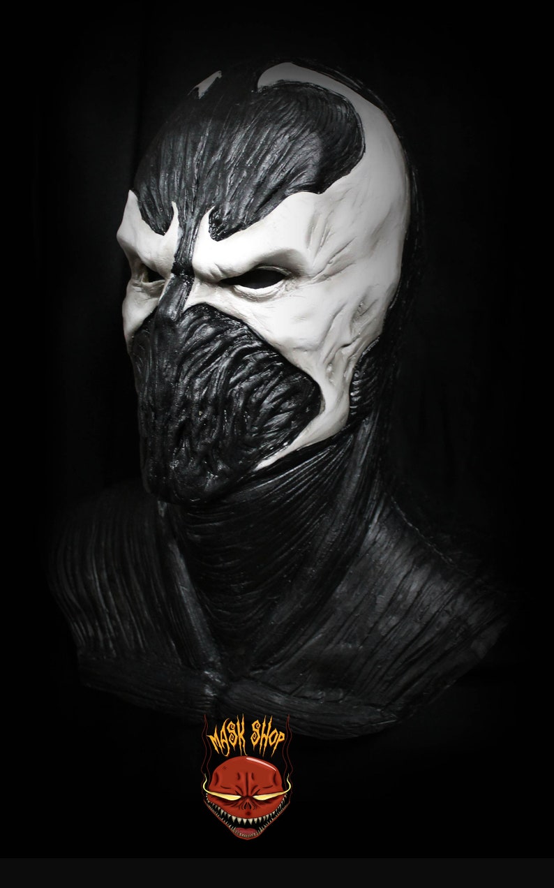 May include: A black and white detailed mask of a character with a large, open mouth and sharp teeth. The mask is made of a textured material and has a realistic appearance. The mask is on a black background.