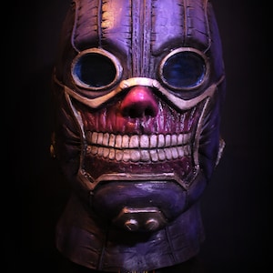 Pu&ograve; includere: Una maschera di teschio viola e oro con un design cucito e occhiali. La maschera ha un ampio sorriso pieno di denti e un naso rosso.