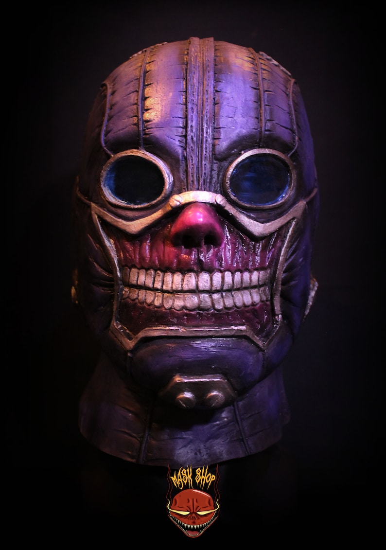 Pu&ograve; includere: Una maschera di teschio viola e oro con un design cucito e occhiali. La maschera ha un ampio sorriso pieno di denti e un naso rosso.