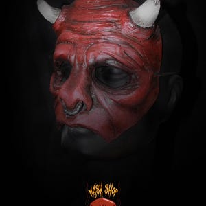 Puede incluir: Una m&aacute;scara de diablo roja con cuernos blancos y un aro en la nariz plateado. La m&aacute;scara tiene una superficie detallada y texturizada, sobre un fondo negro. El logo "Mask Shop" est&aacute; en la parte inferior.