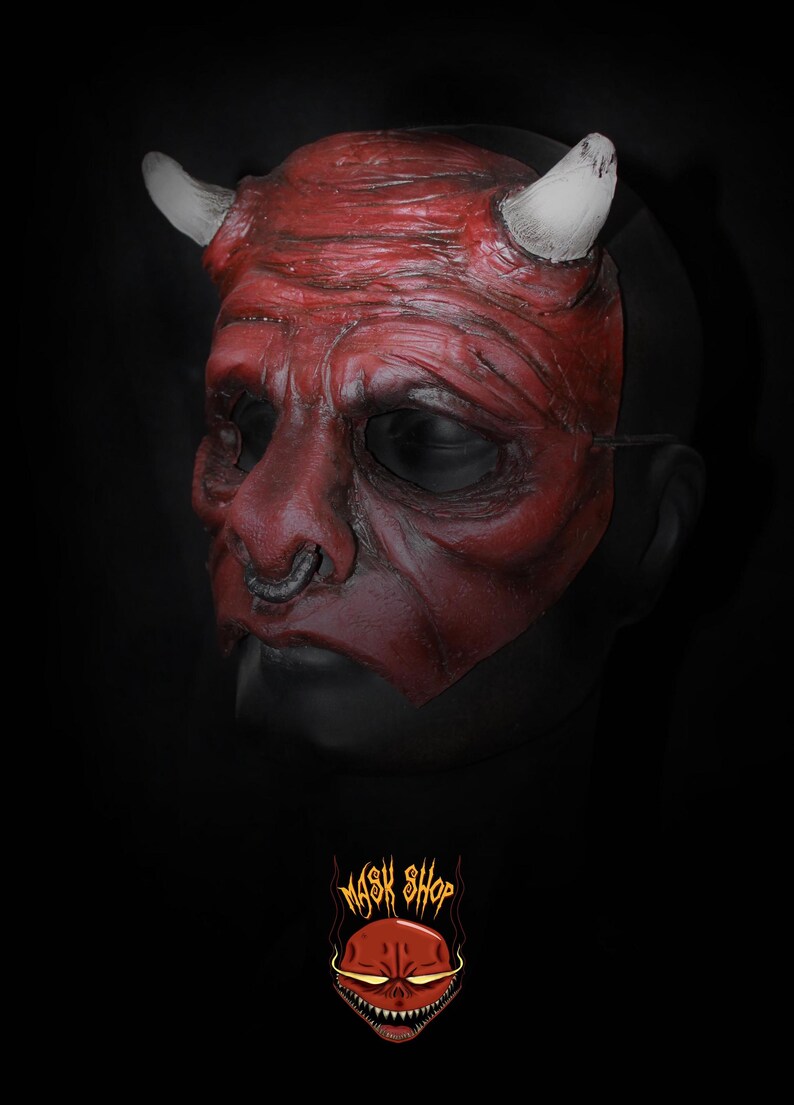 Puede incluir: Una m&aacute;scara de diablo roja con cuernos blancos y un aro en la nariz plateado. La m&aacute;scara tiene una superficie detallada y texturizada, sobre un fondo negro. El logo "Mask Shop" est&aacute; en la parte inferior.