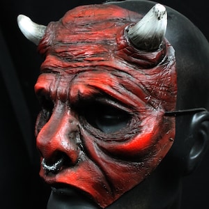 Puede incluir: Una m&aacute;scara de diablo roja con cuernos blancos y un aro en la nariz negro. La m&aacute;scara tiene una apariencia desgastada y texturizada, con arrugas y sombras profundas. La m&aacute;scara est&aacute; montada sobre una cabeza de maniqu&iacute; negra, con fondo negro.