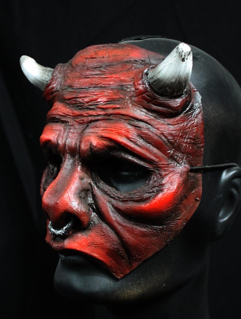 Puede incluir: Una m&aacute;scara de diablo roja con cuernos blancos y un aro en la nariz negro. La m&aacute;scara tiene una apariencia desgastada y texturizada, con arrugas y sombras profundas. La m&aacute;scara est&aacute; montada sobre una cabeza de maniqu&iacute; negra, con fondo negro.