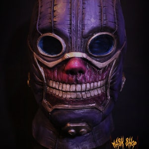 Pu&ograve; includere: Una maschera di teschio viola e oro con un design cucito e occhiali. La maschera ha un ampio sorriso pieno di denti e un naso rosso.