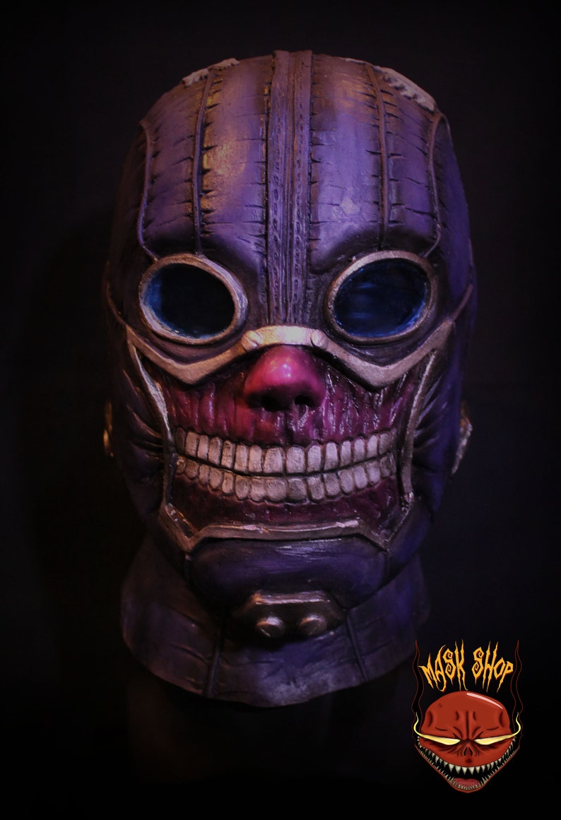 Pu&ograve; includere: Una maschera di teschio viola e oro con un design cucito e occhiali. La maschera ha un ampio sorriso pieno di denti e un naso rosso.