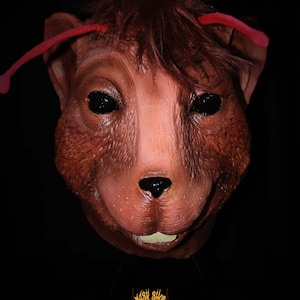 Puede incluir: Una máscara de animal realista de color marrón y rojo con antenas rojas. La máscara tiene ojos negros y una pequeña nariz negra. La boca está abierta y muestra dientes blancos. La máscara está sobre un fondo negro con un logotipo rojo que dice "Mask Shop".
