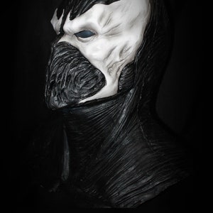 May include: A black and white latex mask of a character with a skull-like face and a black, textured, hooded cape. The mask has a single, large, blue eye.