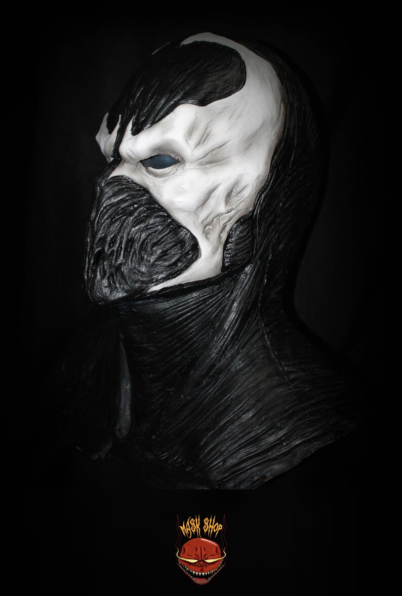 May include: A black and white latex mask of a character with a skull-like face and a black, textured, hooded cape. The mask has a single, large, blue eye.