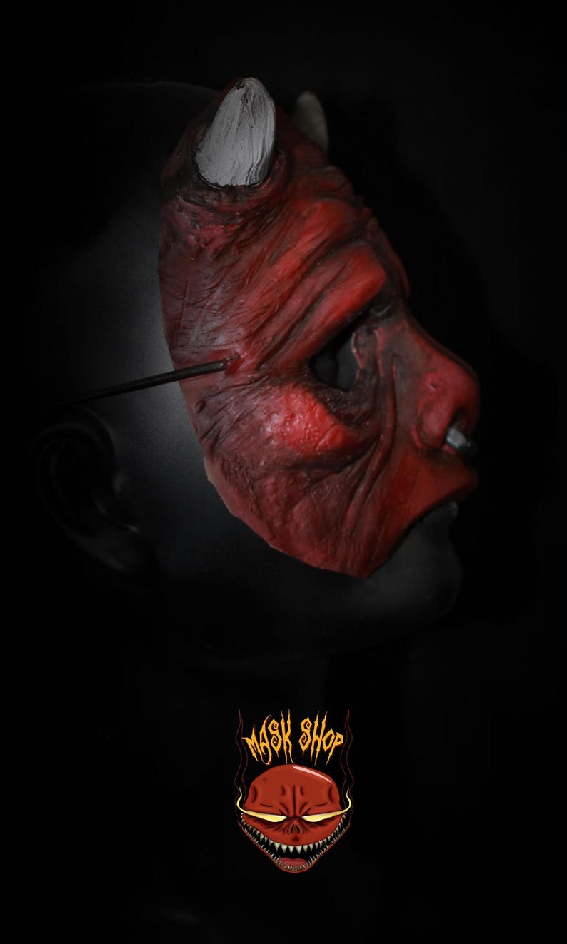 Puede incluir: Una m&aacute;scara de diablo roja con cuernos blancos y una superficie texturizada. La m&aacute;scara tiene una expresi&oacute;n siniestra y est&aacute; dise&ntilde;ada con arrugas y detalles. La imagen incluye el logotipo "MASK SHOP" en la parte inferior.