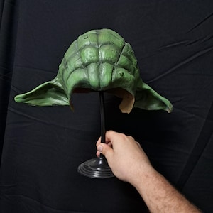 Puede incluir: Máscara verde de Yoda en un soporte negro. La máscara está hecha de un material de goma y tiene una superficie texturizada. A la máscara le faltan los ojos y la boca.