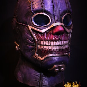 Pu&ograve; includere: Una maschera viola e oro con un designo cucito, occhiali e un ghigno pieno di denti. La maschera ha un naso rosso e un bordo dorato intorno alla bocca.