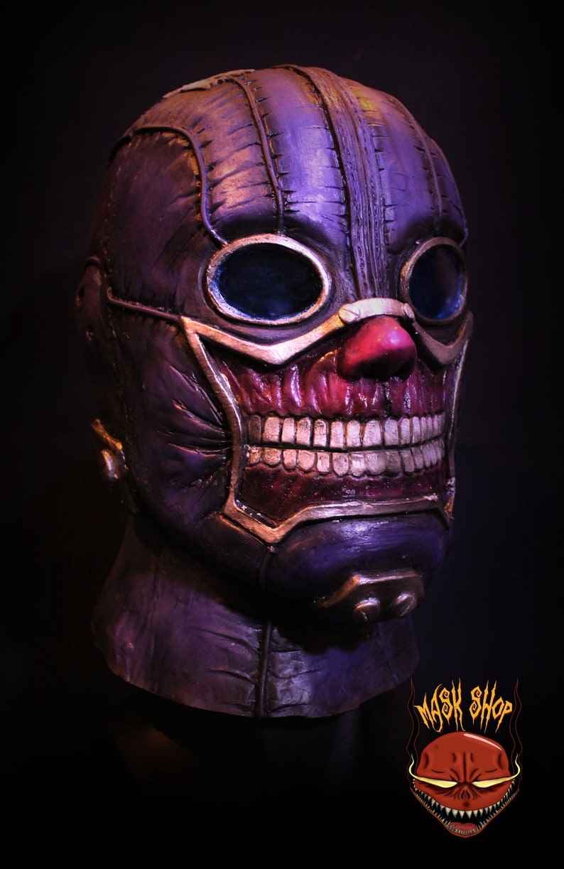 Pu&ograve; includere: Una maschera viola e oro con un designo cucito, occhiali e un ghigno pieno di denti. La maschera ha un naso rosso e un bordo dorato intorno alla bocca.
