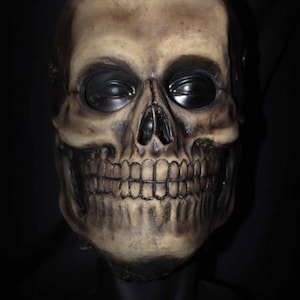 Puede incluir: Una máscara de calavera detallada con una estructura ósea realista y cuencas de ojos negras. La máscara está hecha de un material marrón claro y tiene un aspecto ligeramente desgastado.