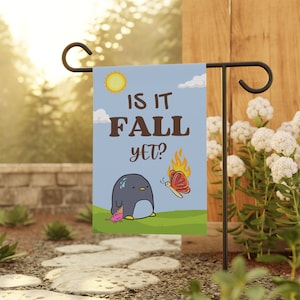 May include: A blue and green garden flag featuring a penguin melting in the sun and a butterfly flying away from a flame. The text on the flag reads "Is it Fall yet?"