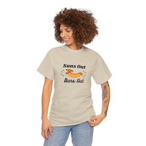 May include: A light beige t-shirt with a graphic of a dachshund wearing a hot dog costume. The text "Suns Out Buns Out" is printed below the graphic.