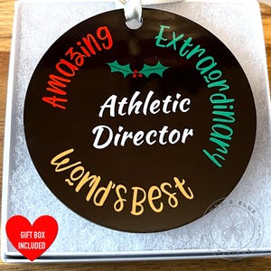 May include: A round, dark brown ornament with the words "Amazing," "Extraordinary," and "World's Best" in red, green, and gold lettering. The text "Athletic Director" is in white. A red heart-shaped sticker reads "Gift Box Included."