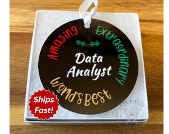 May include: A round black ornament with a white ribbon hanger. The text reads "Amazing" in red, "Extraordinary" in teal, "World's Best" in gold, and "Data Analyst" in white. A small green holly leaf graphic is centered at the top.